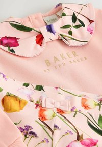 Vestido de algodón rosa con un diseño floral, con un cuello adornado con rosas y un pequeño lazo en la cintura. Logo dorado exhibido.