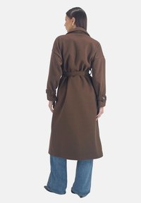 Bruine jas met riem, gemaakt van een gestructureerde stof, met oversized mouwen en zijzakken. Gecombineerd met lichte denim wijde broekspijpen.