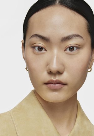 Primo piano di un volto con pelle liscia e capelli scuri e dritti, che indossa orecchini d'oro. Il colletto è di un beige chiaro con dettagli di cucitura ben visibili.