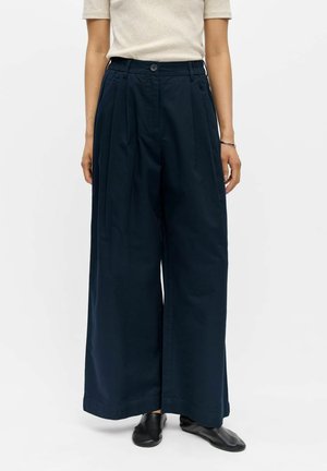 Femme portant un pantalon taille haute large bleu marine, un haut beige à manches courtes, des chaussures noires et un bracelet argenté au poignet droit.