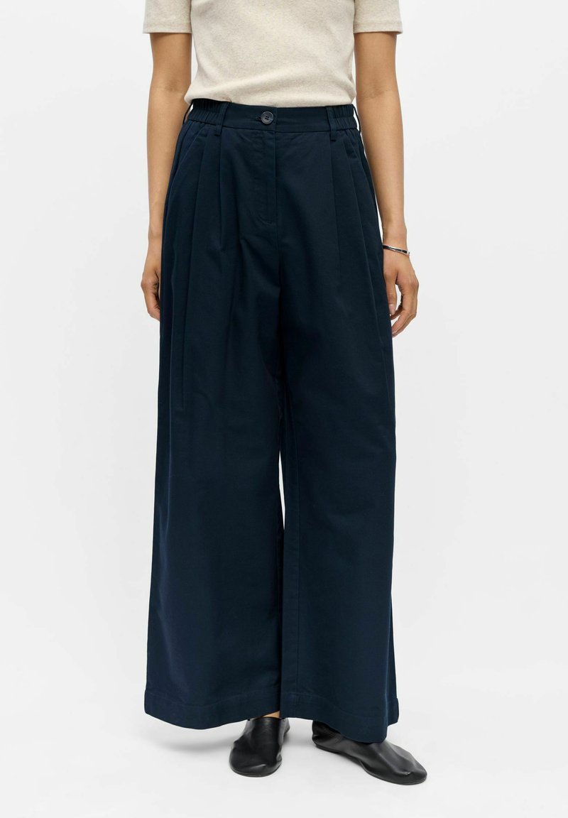 Femme portant un pantalon taille haute large bleu marine, un haut beige à manches courtes, des chaussures noires et un bracelet argenté au poignet droit.