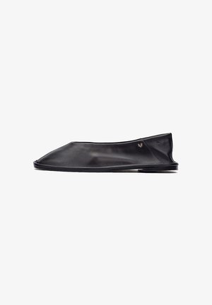 Zapato plano minimalista de cuero negro con un tacón bajo y pequeño logotipo metálico cerca del talón, mostrado de perfil sobre un fondo blanco.