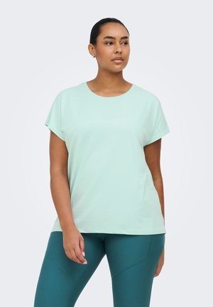 Vrouw met donker haar, gekleed in een lichtblauw T-shirt met korte mouwen en groenblauwe legging, staand met een neutrale uitdrukking tegen een effen achtergrond.