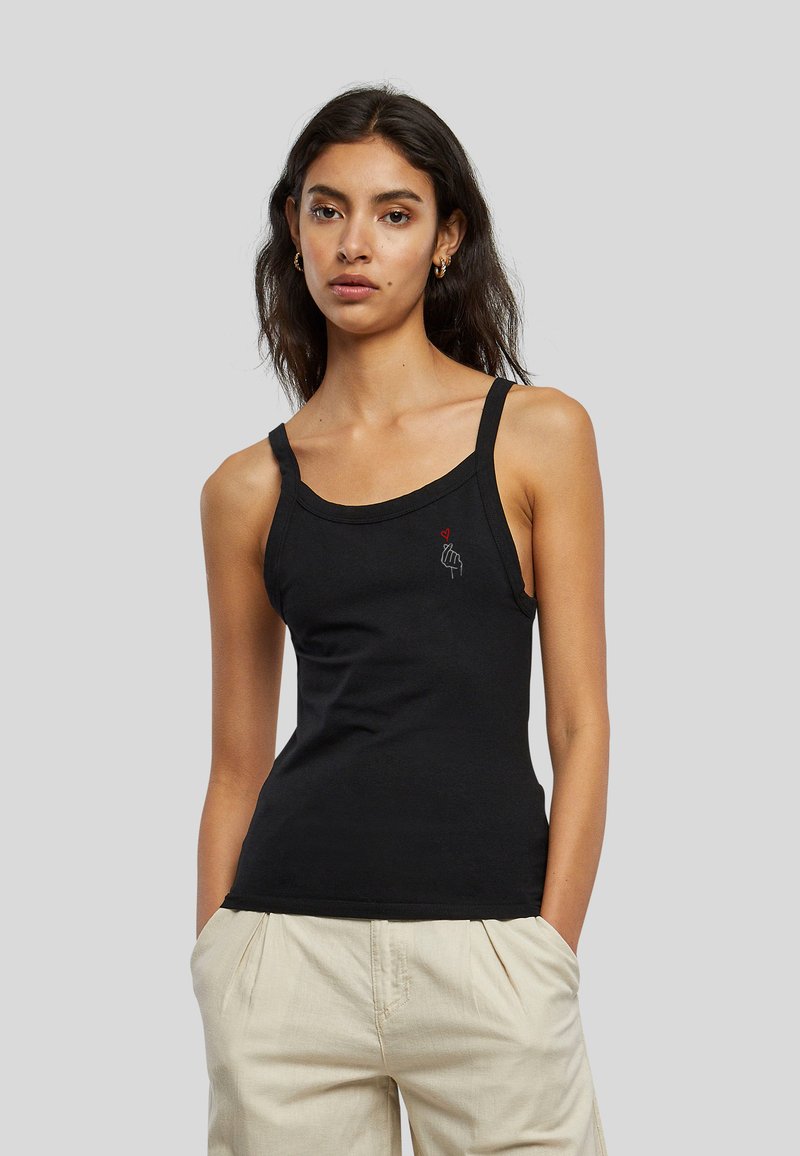 Femme aux longs cheveux foncés porte un débardeur noir avec un petit motif de main et cœur, pantalon beige, debout devant un fond gris.