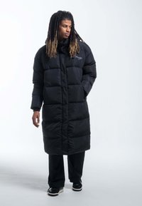 Lungo cappotto imbottito nero con design trapuntato, colletto alto e zip frontale. Completato da pantaloni e scarpe nere.