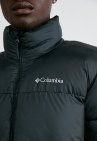 Sort pufferjakke med høj krave, lynlåslukning foran og Columbia-logo på brystet. Glat tekstur med et subtilt mønster.