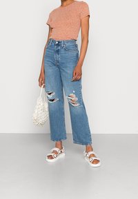 Oranžový top s krátkým rukávem a bílými tečkami, vysoké roztrhané modré džíny a bílé sandály. Dokončeno texturovanou bílou taškou.