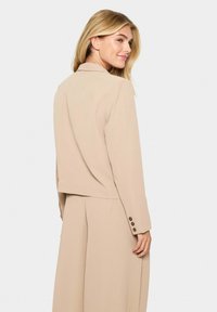 Beige zkrácený blazer s přizpůsobeným střihem, klopovým límcem a knoflíkovými manžetami. Hladká látka a čisté linie doplňují minimalistický styl.