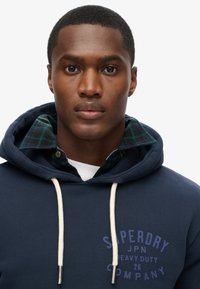 Felpa blu navy con lacci bianchi e logo viola, indossata sopra una camicia a quadri verde e nera. Texture del tessuto liscia, design casual.
