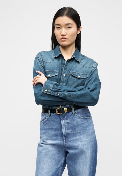 Νεαρή γυναίκα με ίσια μαύρα μαλλιά, ντυμένη με μπλε τζιν πουκάμισο και ψηλόμεσα τζιν, στέκεται με σταυρωμένα χέρια, με ουδέτερη έκφραση.