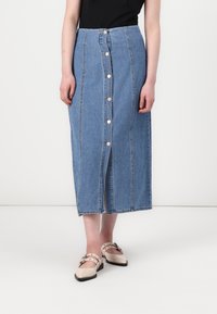 Jupe en denim bleu clair, mi-longue, avec une fermeture à boutons sur le devant, des coutures verticales et un ourlet effiloché. Associée à des chaussures beiges.