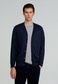 Marinblå strikket cardigan med v-hals, frontknapper og ribbede manchetter, båret over en grå t-shirt. Glat tekstur, enkelt design.
