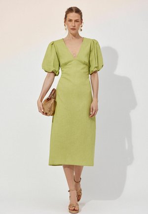 Frau trägt ein hellgrünes Midikleid mit Puffärmeln, hält eine kleine braune Handtasche und trägt beige Riemchenabsätze, steht an einer weißen Wand.
