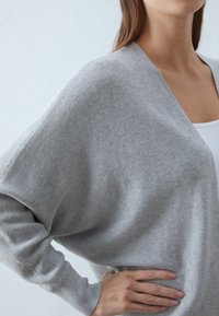 Šedý žebrovaný cardigan se širokým výstřihem, spadlými rameny a uvolněným střihem. Měkká textura s dlouhými rukávy, nošený přes bílý top.