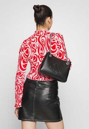 Zwarte leren handtas met reliëflogo, zilveren kettingband en gestructureerde vorm. Model draagt een rood-witte gedraaide top en een zwarte leren rok.