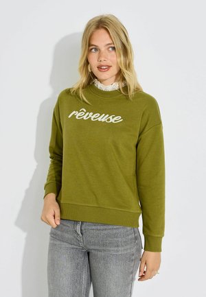 Mujer rubia con sudadera verde oliva con texto "rêveuse" y vaqueros grises, de pie contra un fondo blanco mirando a la derecha.