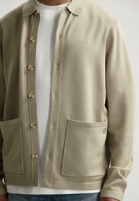 Vaaleanoliivinvihreä neuletakki, joka on valmistettu pehmeästä neuloksesta. Siinä on napitus edessä, kaksi suurta taskua ja kauluskainalo.