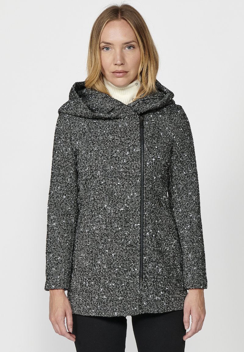 Čierno-biely škvrnitý kabát s veľkou kapucňou, zipsovým zapínaním, prispôsobeným strihom a dlhými rukávmi. Mäkky materiál, dĺžka do bokov.
