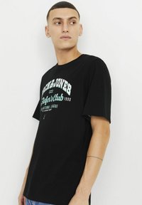 Sort bomuld T-shirt med korte ærmer, prydet med hvid og teal grafisk print, der står "Jack & Jones" og "Buffer's Club 1990."