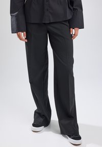 Black wide-leg trousers made of smooth fabric, featuring a tailored fit and front crease. Worn with black and white sneakers.