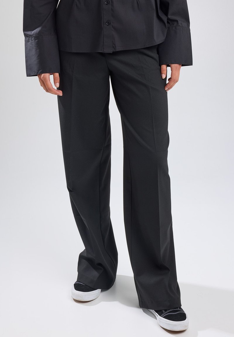 Black wide-leg trousers made of smooth fabric, featuring a tailored fit and front crease. Worn with black and white sneakers.