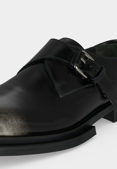 Μαύρο δερμάτινο παπούτσι monk strap με ασημένια αγκράφα και ελαφρώς φθαρμένη μύτη, εμφανίζεται από κοντινή λοξή πλευρική όψη.