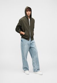 Νεαρός άνδρας που φοράει ελιάς ζακέτα με φερμουάρ και τα χέρια στις τσέπες, ανοιχτό μπλε φαρδιά τζιν και λευκά αθλητικά παπούτσια, στέκεται μπροστά σε λευκό φόντο.