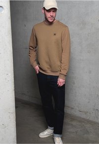 Brauner Sweatshirt mit Textur und Logo, kombiniert mit dunkelblauen Jeans und weißen Sneakers. Mann lehnt an einer Betonwand.