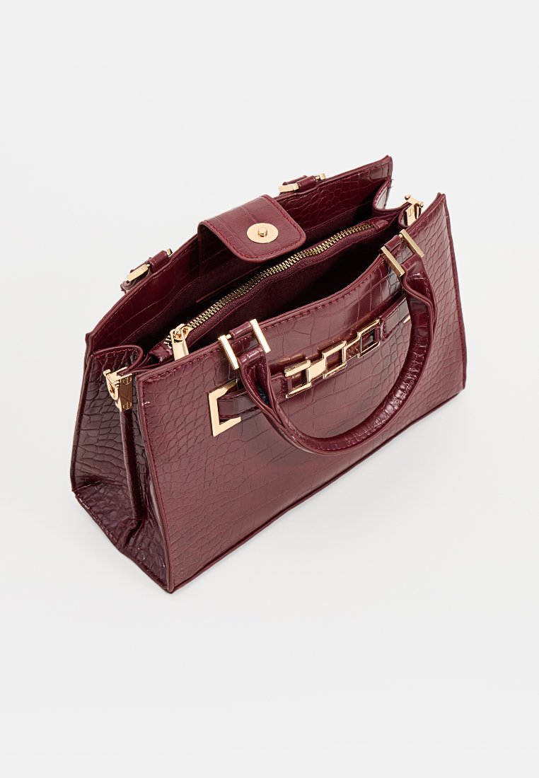 Sac à main en cuir d'alligator bordeaux avec quincaillerie dorée, forme structurée, doubles poignées et fermeture éclair sécurisée. Poche intérieure visible.