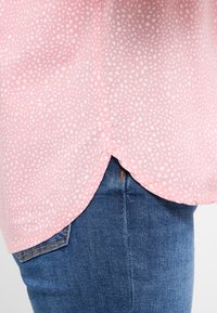 Helles rosa Hemd mit einem weißen gesprenkelten Muster, mit abgerundetem Saum und weichem Stoff, getragen mit blauen Jeans.