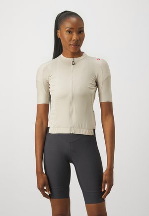 Beige short-sleeve cycling jersey with a front zipper, featuring a textured fabric and a small logo on the shoulder. Black cycling shorts.