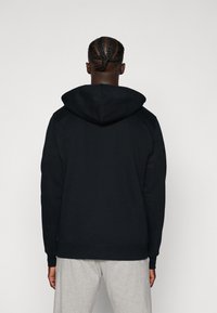 Schwarzer Kapuzenpullover mit lockerer Passform, gerippten Bündchen und Saum, von hinten gezeigt, kombiniert mit hellgrauen Hosen.