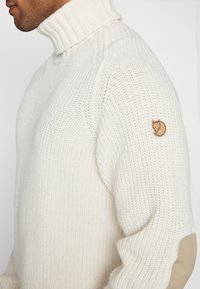 Maglione bianco a collo alto in maglia con colletto a coste, dotato di una toppa con logo marrone sulla spalla sinistra e una superficie testurizzata.