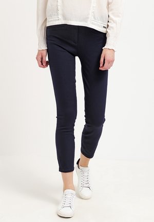 Femme portant un pantalon slim à chevilles bleu marine, un chemisier blanc à manches longues et des baskets blanches, marchant sur un fond blanc uni.