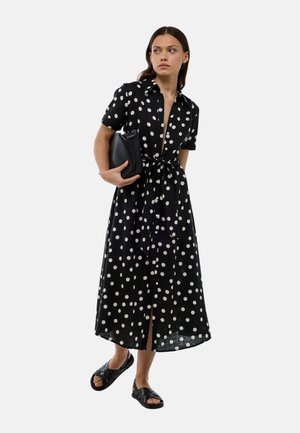 Robe chemise noire à pois blancs avec des manches courtes, une ceinture à la taille et un devant à boutons. Portée avec des sandales noires et tenant un sac noir.