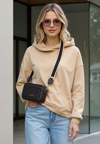 Haut à capuche beige avec poche avant, associé à un jean bleu clair. Un petit sac en bandoulière noir présente une surface texturée et une sangle ajustable.