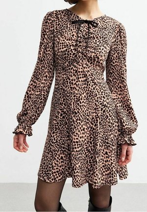 Femme portant une robe imprimé léopard à manches longues, longueur genoux, avec un ruban noir noué au niveau de l'encolure et des poignets volantés.