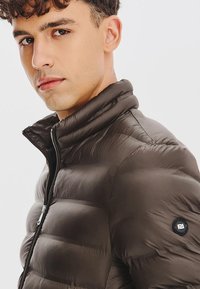 Brązowa kurtka puchowa z wysokim kołnierzem, pikowanym wzorem, przednim zamkiem i okrągłą naszywką z logo na lewym rękawie.