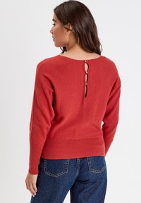 Suéter rojo de punto con un detalle de ojal en la espalda, que presenta dobladillos y mangas acanaladas. La tela parece suave y ligeramente texturizada. Combinado con jeans oscuros.