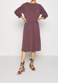 Mauve, knälång klänning med trekvartslånga puffärmar och resår i midjan. Kombinerad med svarta sandaler med klack och remmar. Slät textur.