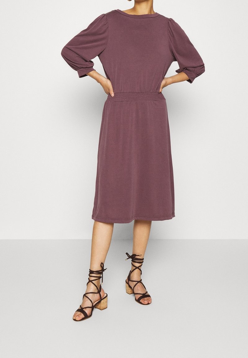 Mauve, knälång klänning med trekvartslånga puffärmar och resår i midjan. Kombinerad med svarta sandaler med klack och remmar. Slät textur.