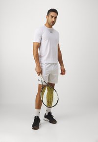 Camiseta atlética blanca con un logo sutil, combinada con pantalones cortos blancos. Sostiene una raqueta de tenis con un patrón de cuerdas amarillas y zapatillas deportivas negras.