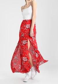 Jupe longue fleurie rouge avec des fentes latérales, en tissu léger. Associée à un débardeur blanc et des baskets blanches.