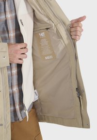 Beige vandtæt jakke med en tekstureret indre foring, lynlås lukning og knappet front. Inkluderer en plejeetiket indeni. Har en hætte.