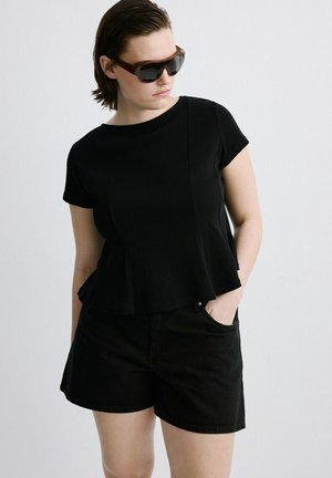 Osoba oblečená v čiernom krátkom rukáve a čiernych šortkách, s tmavými slnečnými okuliarmi, pózujúca s jednou rukou v vrecku na jednoliatom pozadí.