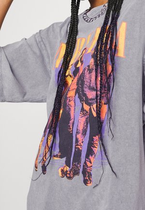 Persoon met lange vlechten die een grijs Nirvana grafisch T-shirt draagt met een oranje en paarse bandprint en een zilveren ketting.