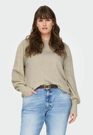 Pull en maille vert olive clair avec des manches bouffantes, présentant des motifs texturés découpés. Associé à un jean bleu et une ceinture marron à double boucle.