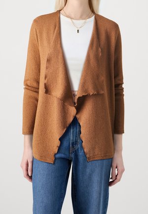 Frau trägt einen braunen offenen Cardigan über einem weißen gerippten Crop-Top und blauen Jeans, mit geschichteten goldenen Halsketten.