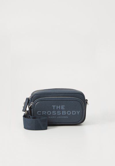 Compact navy blauwe leren crossbody tas met twee ritscompartimenten en een verstelbare band, gemerkt "THE CROSSBODY MARC JACOBS" aan de voorkant.