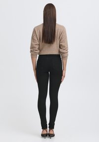 Juodos lieknos džinsinės kelnės su aukštu liemeniu ir galiniais kišenėmis, derinamos su smėlio spalvos megztiniu, turinčiu laisvą siluetą ir raukiniais.